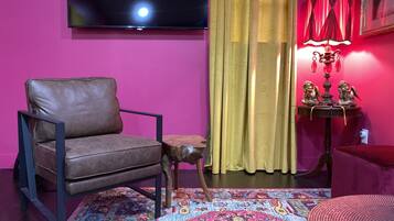 Una sala de estar con un sillón de cuero marrón, una mesita de madera, un tapete colorido, un televisor montado en la pared, una lámpara roja y una cortina.