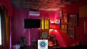 Une pièce avec un mur rouge, un téléviseur à écran plat fixé au mur, une table avec une lampe et une chaise. Il y a une plante sur le sol et une pancarte avec le texte « SANFORD SHORT TERM SINCE 2021 ».