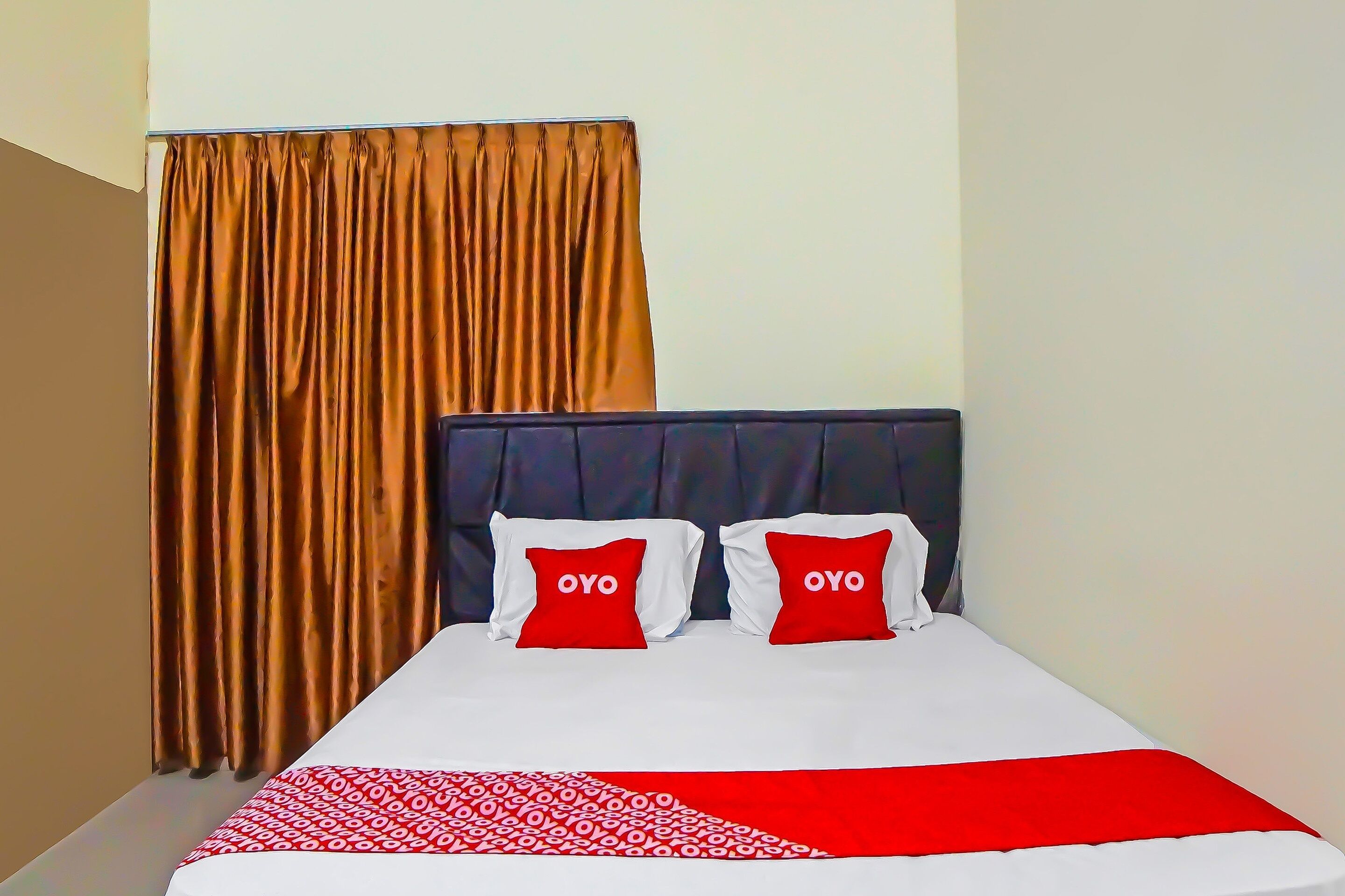A hotel room with a bed, two red pillows with the word "OYO" on them, a patterned throw blanket, and a curtain.