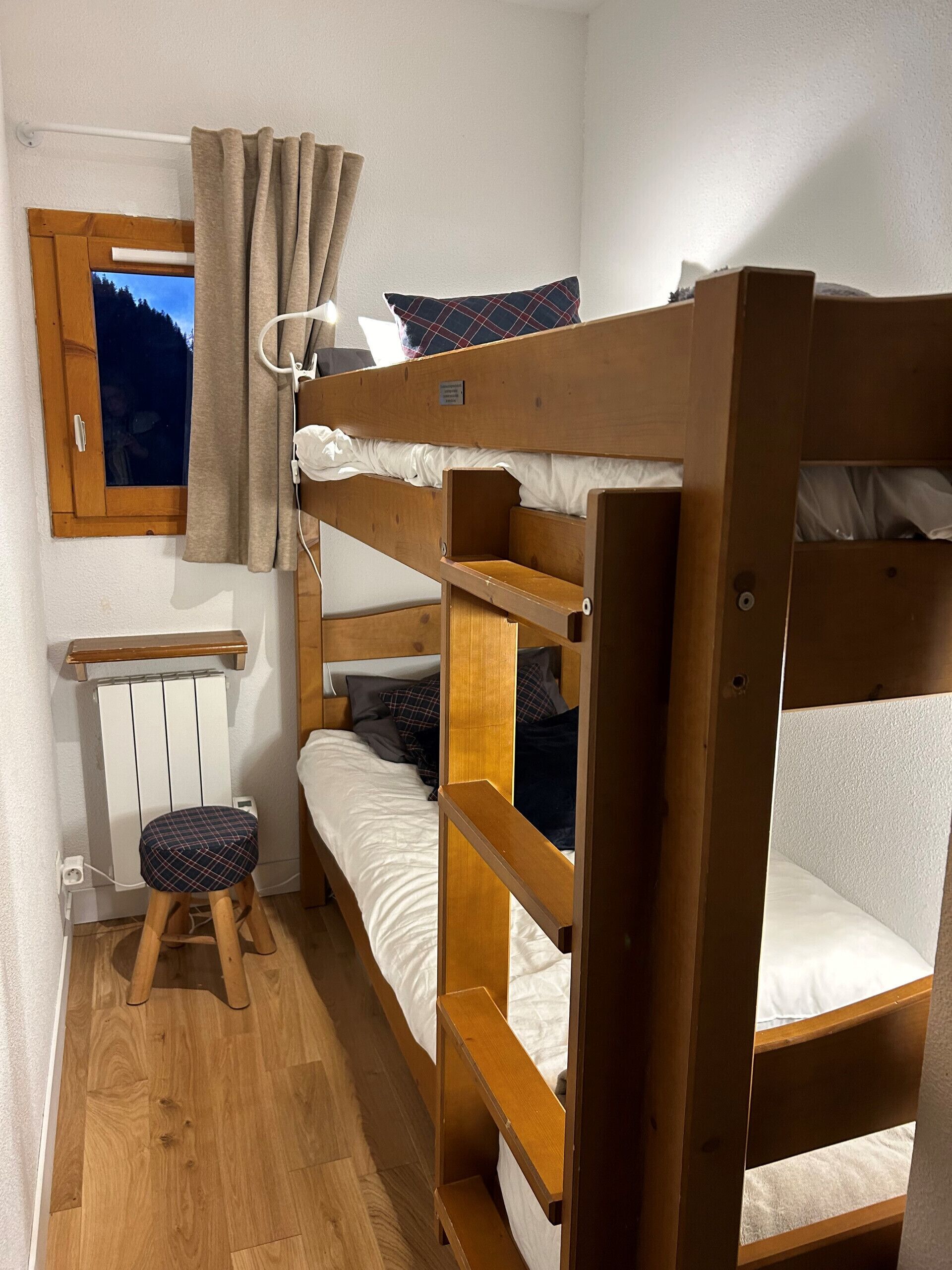 Une chambre avec un lit superposé, un plancher en bois, un petit tabouret, une fenêtre avec des rideaux et un radiateur.