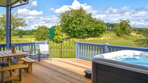 Une terrasse en bois avec un jacuzzi et une table entourée de chaises, offrant une vue sur un paysage verdoyant.