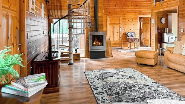Un intérieur spacieux de chalet en bois avec une cheminée centrale, un escalier en colimaçon et un coin salon confortable comprenant un canapé et une table basse.