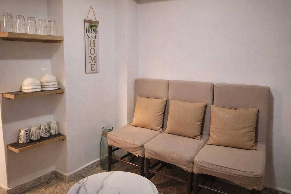 Une pièce aux couleurs neutres, avec un canapé beige, deux étagères en bois ornées de verrerie et d’objets décoratifs, et un panneau mural sur lequel on peut lire : « Chez soi, on est bien ».