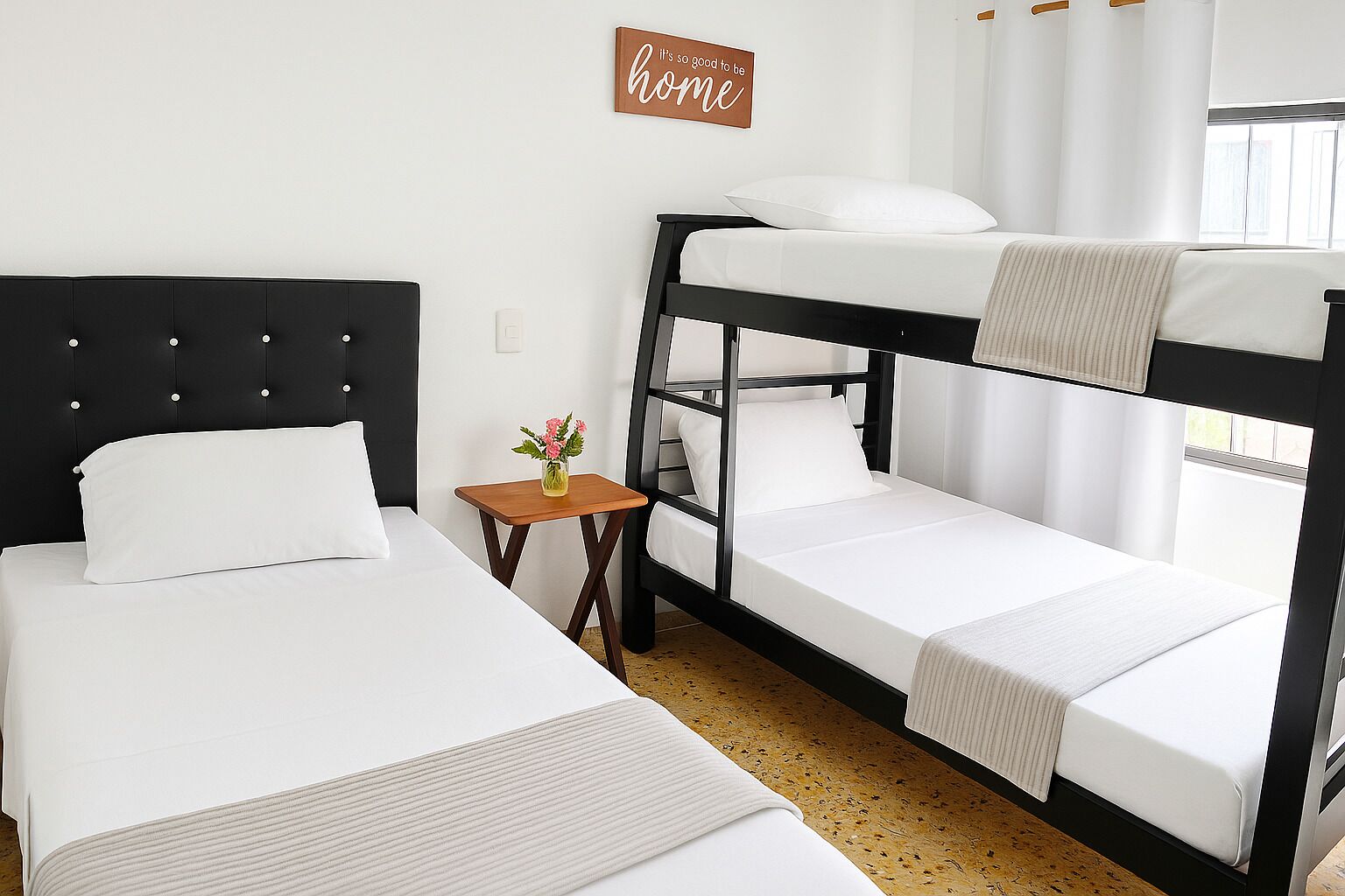 A room with two beds, one with a headboard and the other with a bunk bed. There is a small table with a vase of flowers, and a sign on the wall that reads "It's so good to be home."