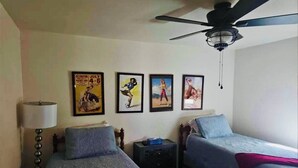 Une chambre avec deux lits simples, un ventilateur de plafond, une table de chevet, une lampe et quatre affiches encadrées accrochées au mur.