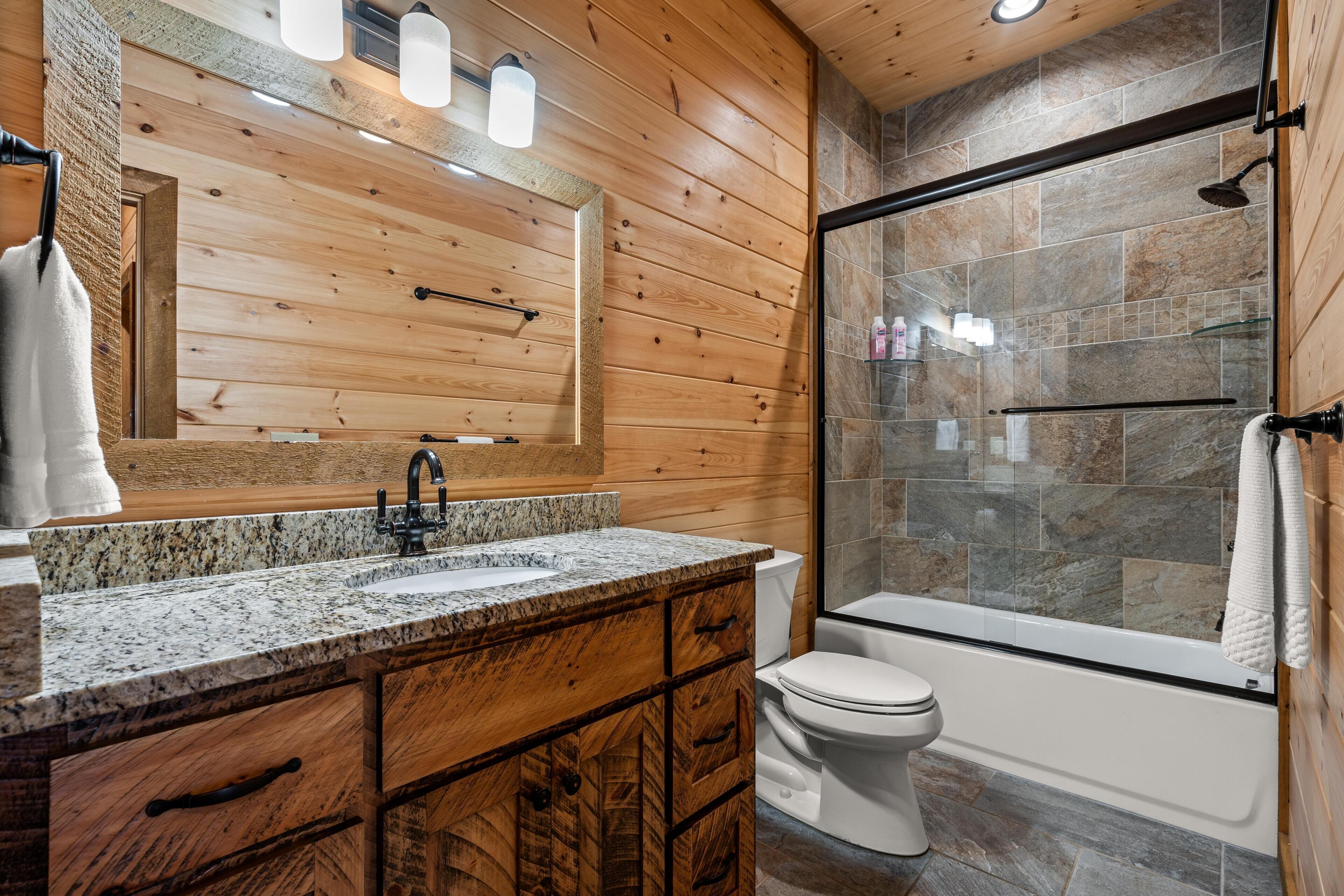 A bathroom with a wooden vanity, a granite countertop, a white toilet, and a glass-enclosed shower area.