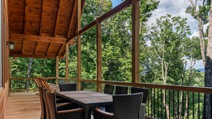 Una terraza de madera con mesa y sillas, rodeada por una barandilla y vistas a árboles.