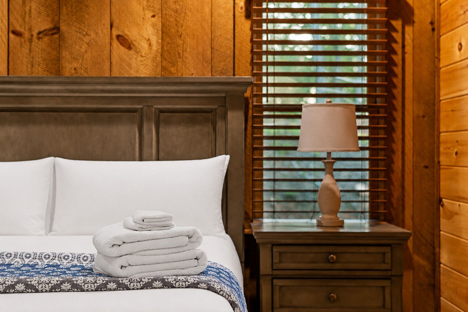 A neatly made bed with white linens and a folded towel stack, a wooden nightstand with a lamp, and a window with blinds.