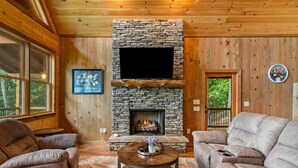 A cozy living room with a stone fireplace, leather sofas, and a flat-screen TV. - Spacious mountain cabin w/mountain views, hot tub & game room! (Cherry Log)
