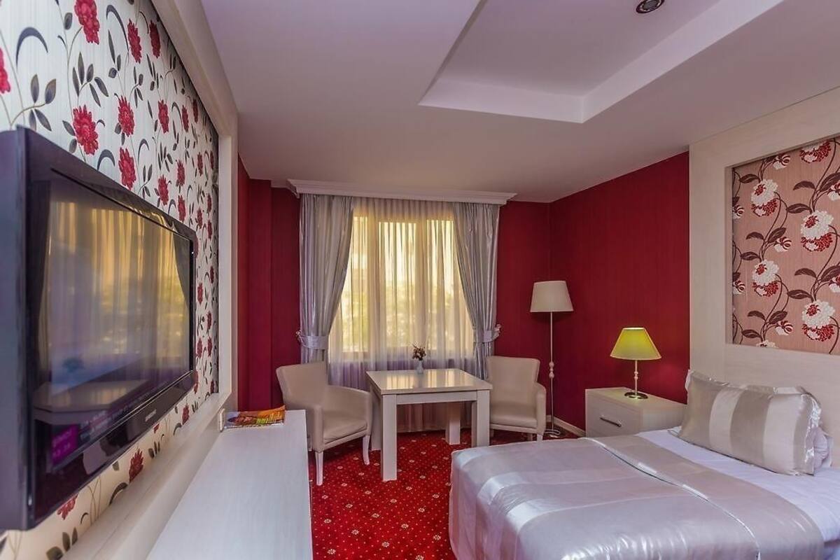 A hotel room with a TV, a bed, a desk, two chairs, a lamp, and a window with curtains.