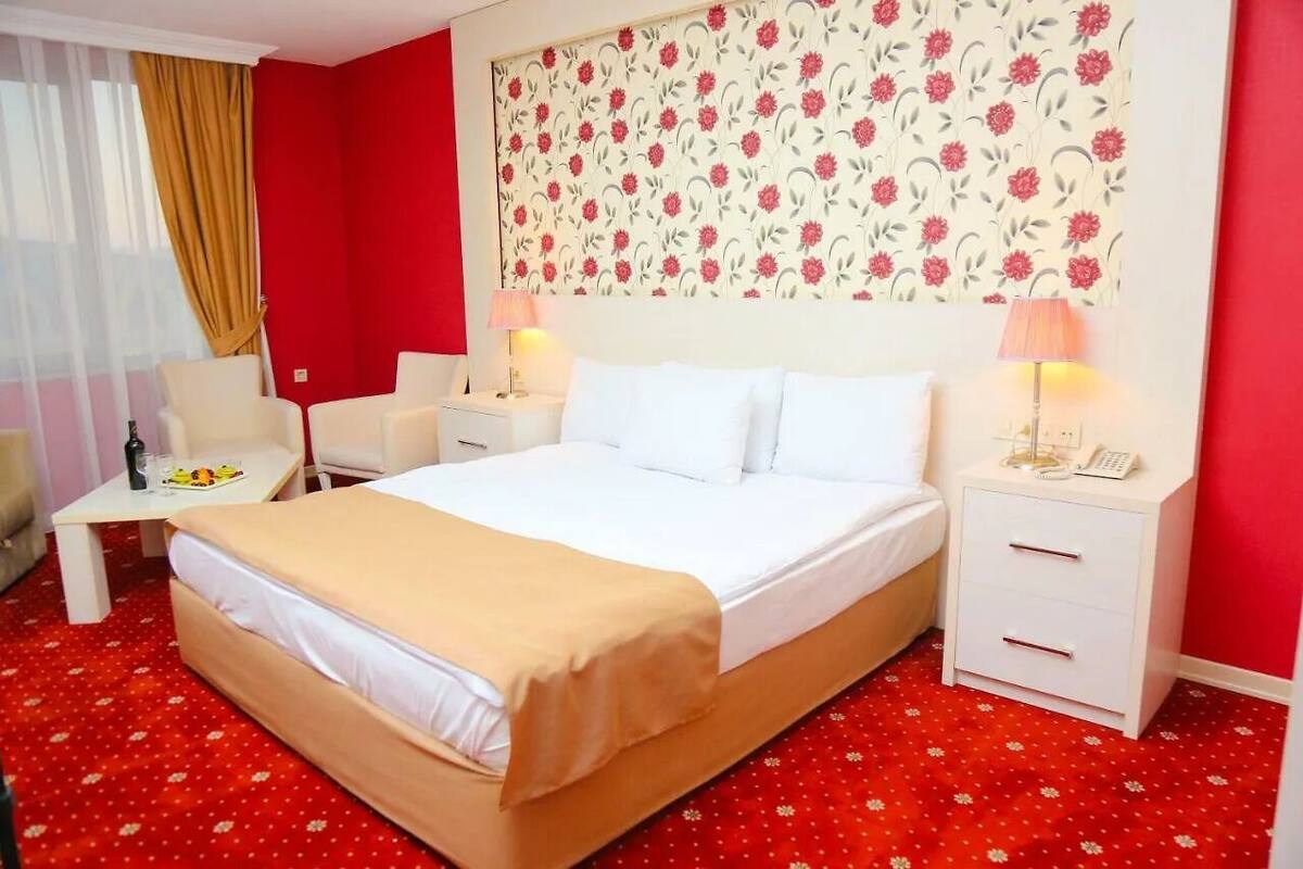 A hotel room with a large bed, two bedside tables, a chair, a small table with a bottle and glasses, and a floral-patterned headboard.