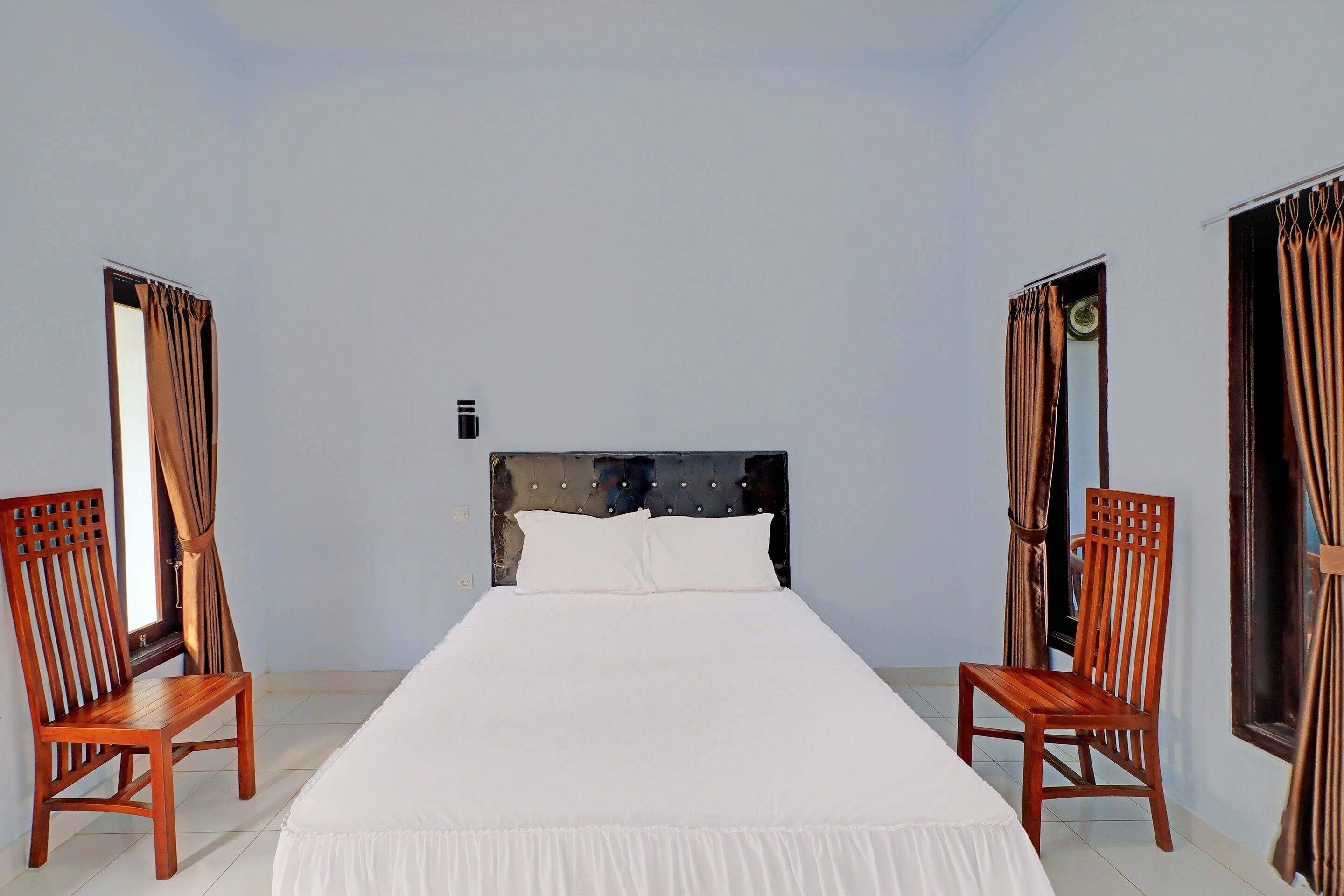 Een netjes ingerichte hotelkamer met een groot bed, twee houten stoelen en gordijnen voor de ramen.