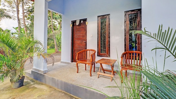 A covered porch with a wooden table and chairs, a potted plant, and a white wall with a blue door. - Hotel O Mustika (Negara)