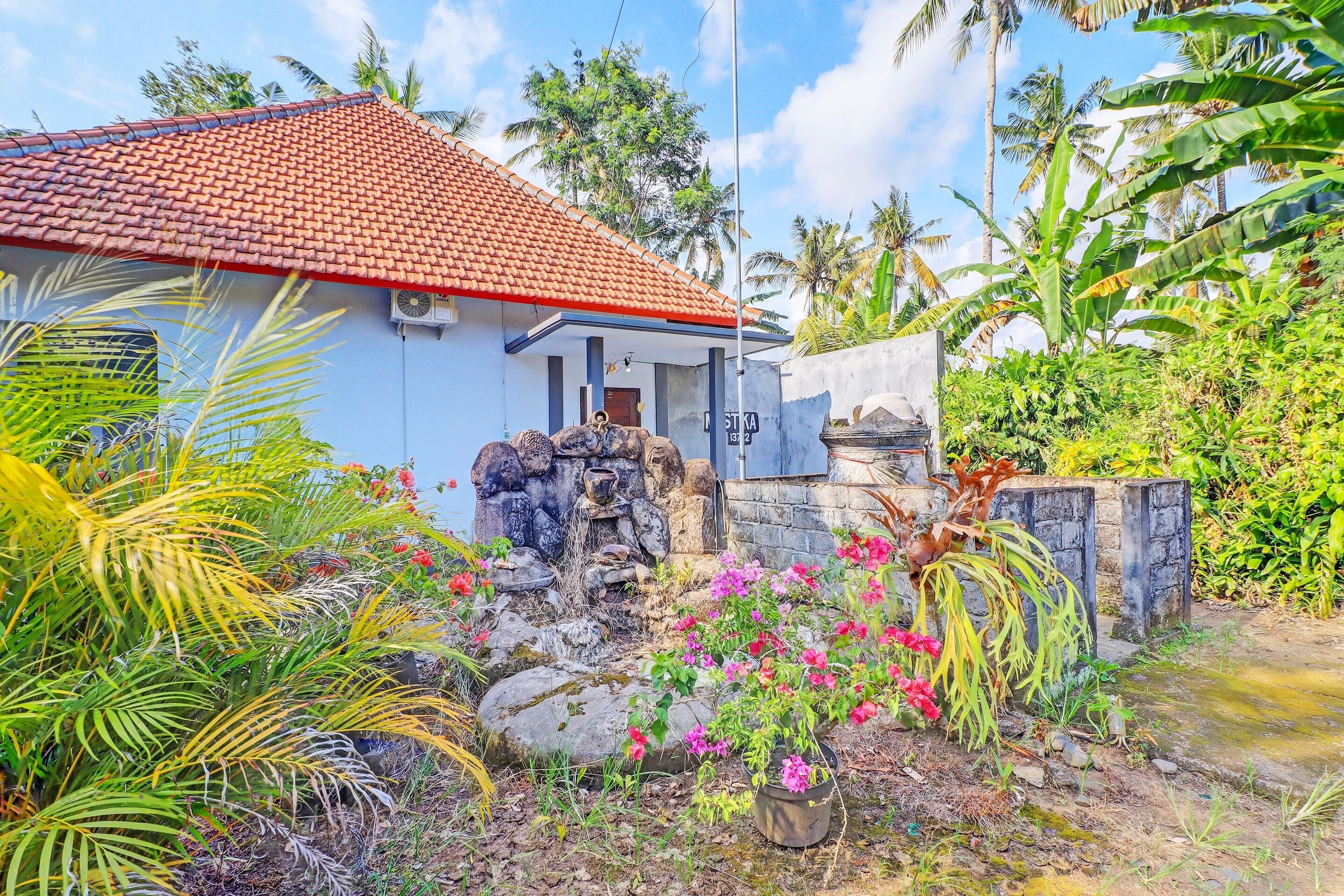 Uma casa com telhado de telhas vermelhas, um elemento decorativo de água em pedra e vegetação tropical exuberante.
