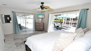 A bedroom with a bed, a dresser, a TV, and a view of an outdoor pool area. - Coral Sunshine - large south terrace with pool/spa (Cape Coral)