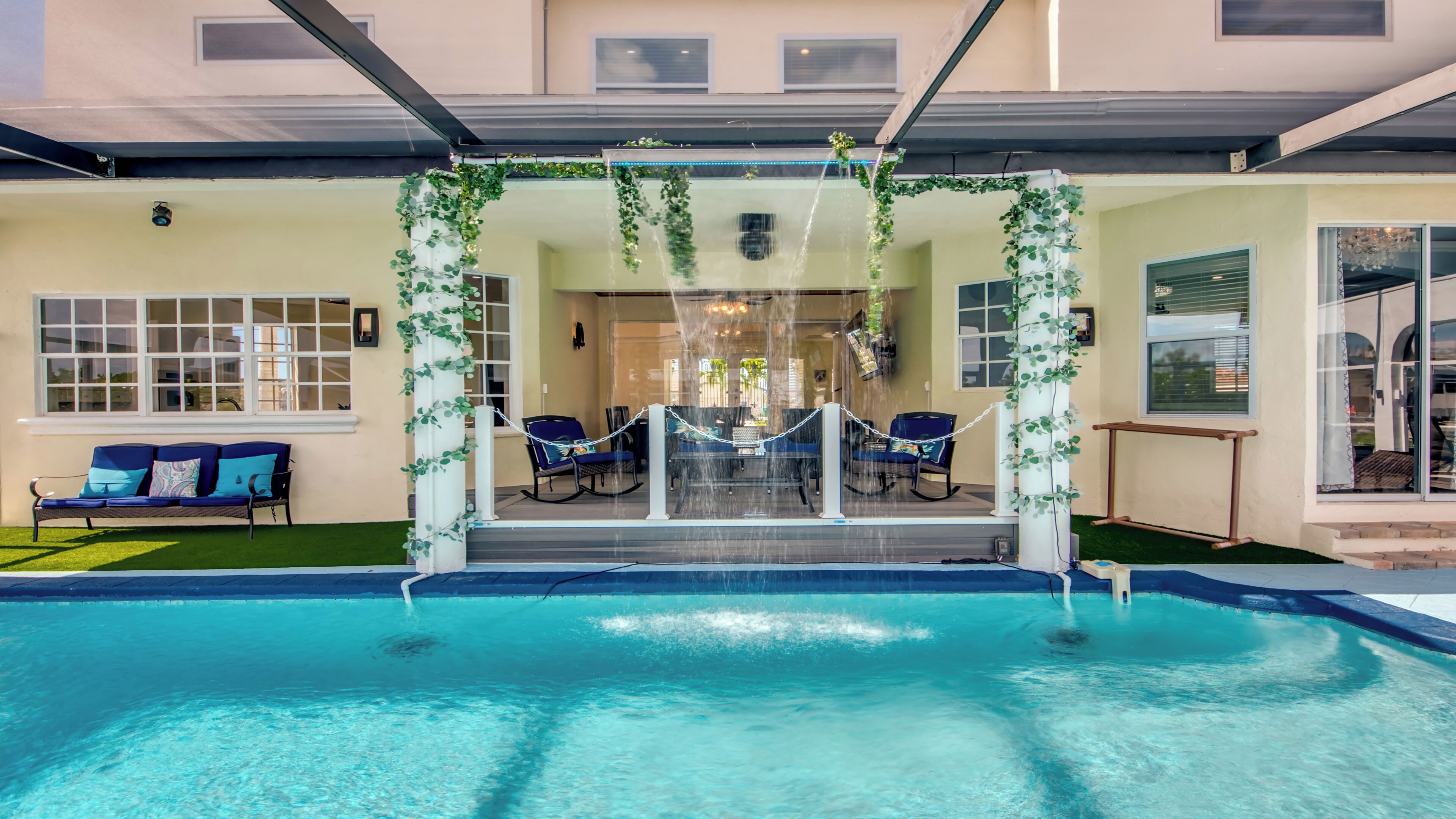 Un espace piscine comprenant un coin salon, une partie couverte avec une table et des chaises, et une vue sur l’intérieur à travers une porte vitrée.