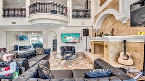 A spacious living area with a black leather sofa, a glass coffee table, a flat-screen TV, a fireplace with a guitar, and a balcony with a wrought iron railing. - Golden Globe - luxury on the canal with pool/spa (Cape Coral)