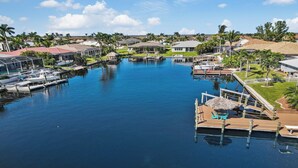 A marina with boats docked, a dock with a thatched umbrella, and houses with yards and palm trees in the background. - Lupita, boat lift, dock, heated south facing pool (Cape Coral)