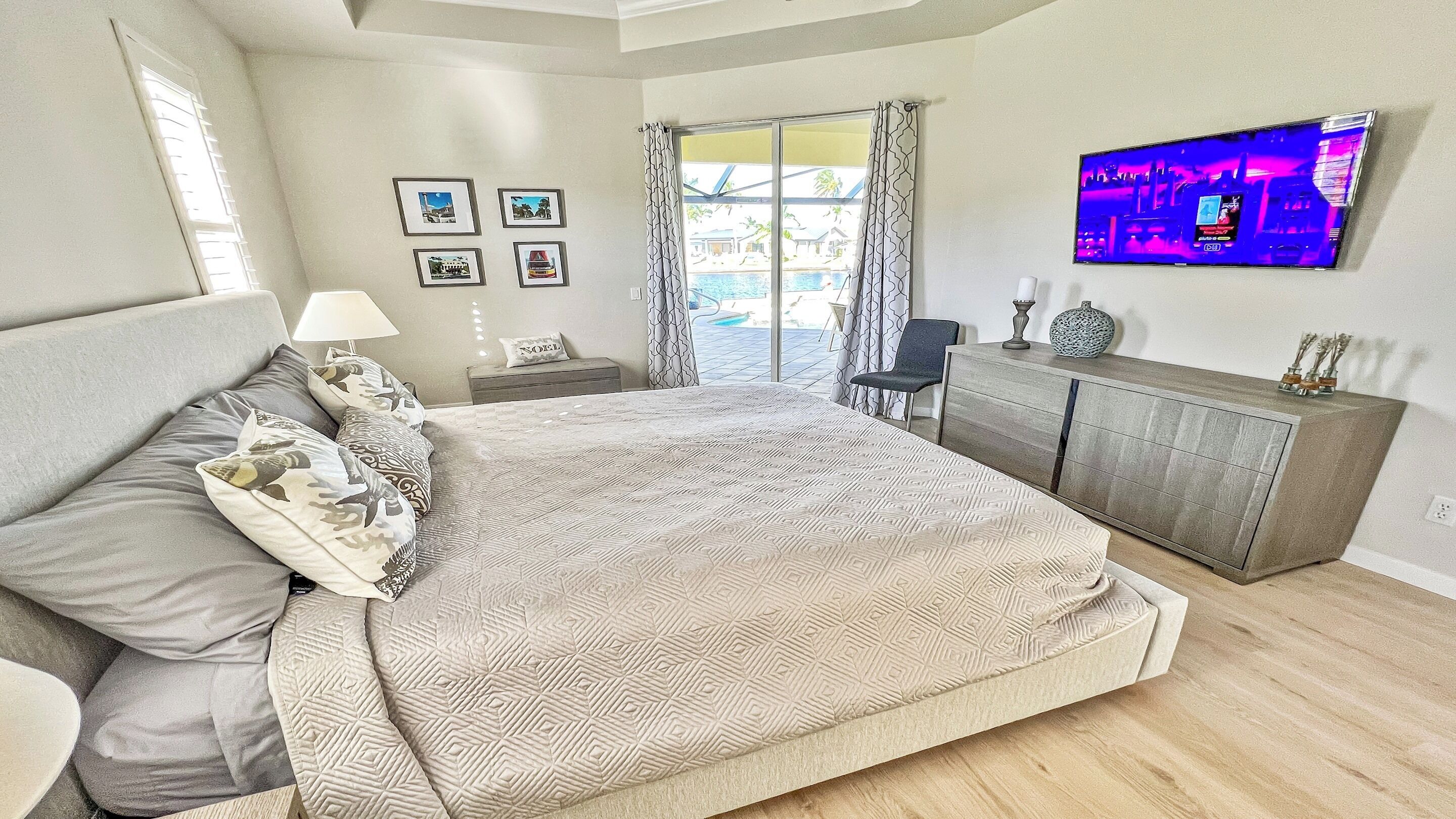 A bedroom with a large bed, a TV mounted on the wall, and a view of a pool through the window.