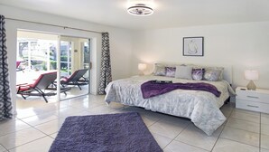 Une chambre à coucher avec un lit, une table de chevet, une lampe, un plaid violet et une vue sur un espace extérieur aménagé avec des chaises et une table.