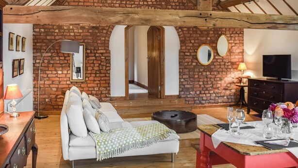 Une pièce chaleureuse avec un mur en briques, des poutres apparentes, un canapé, une table à manger dressée avec des verres et un téléviseur.