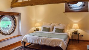 Une chambre avec un plafond en bois, une grande fenêtre ronde, un lit recouvert de linge de maison blanc, deux tables de chevet avec des lampes, et une vue sur un champ verdoyant où paissent des moutons.