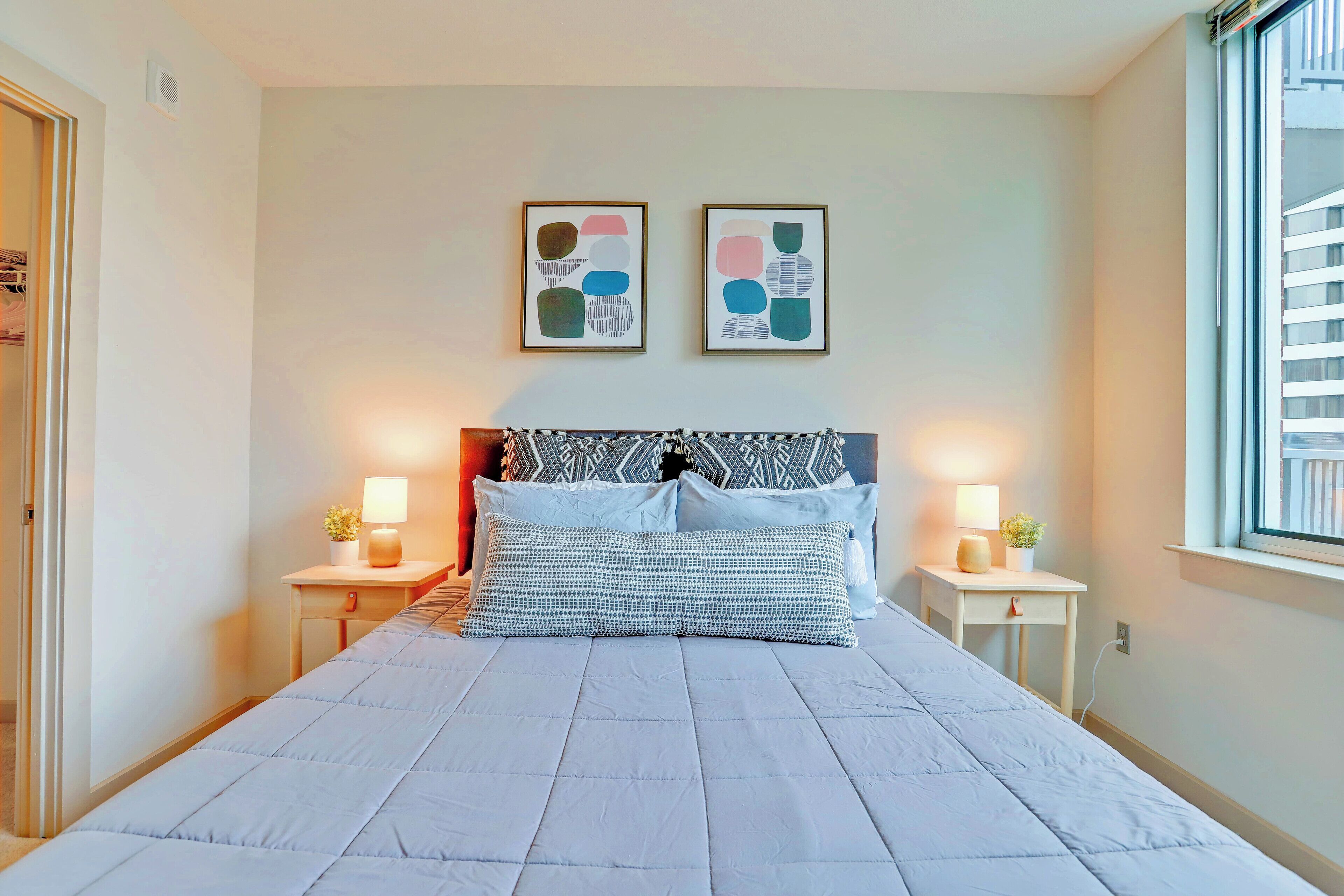 Una cama bien hecha con un almohadón estampado, dos mesitas de noche con lámparas y una obra de arte abstracta en la pared.