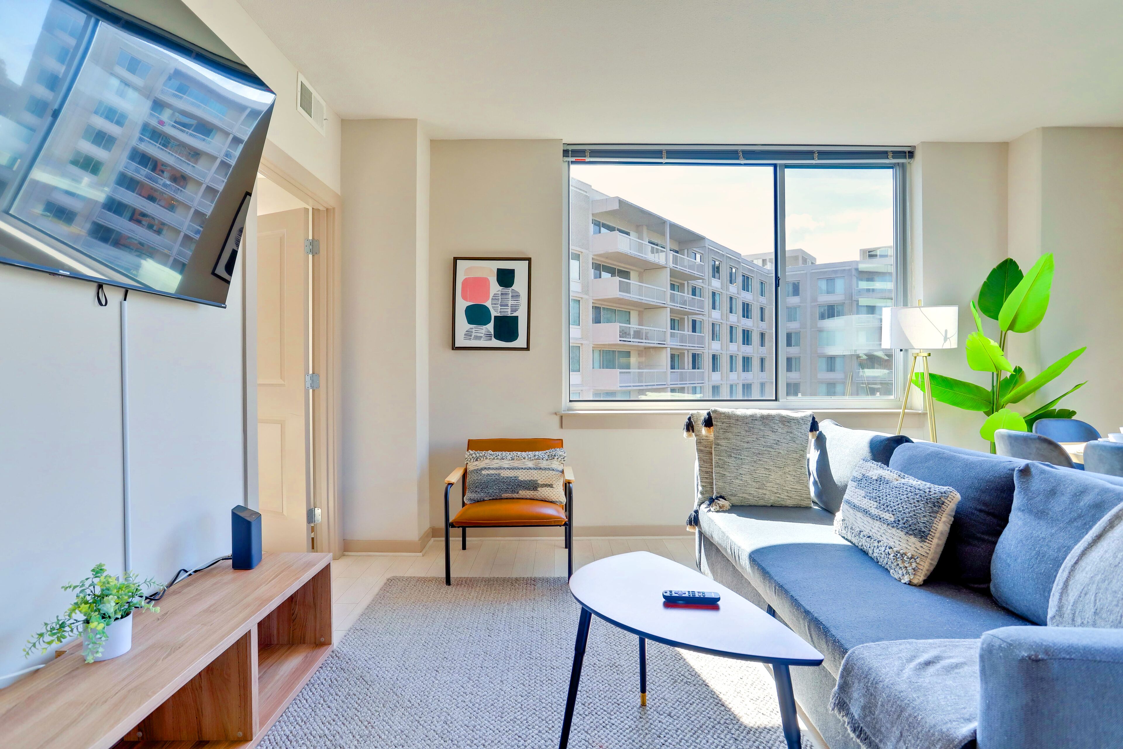 Una sala de estar moderna con un sofá, una mesa de centro, un televisor montado en la pared, una silla y un ventanal con vistas a edificios.