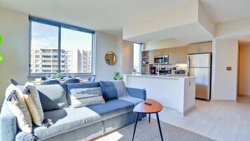 A modern apartment with a grey sofa, a small round table, and a kitchen area with a fridge and microwave.