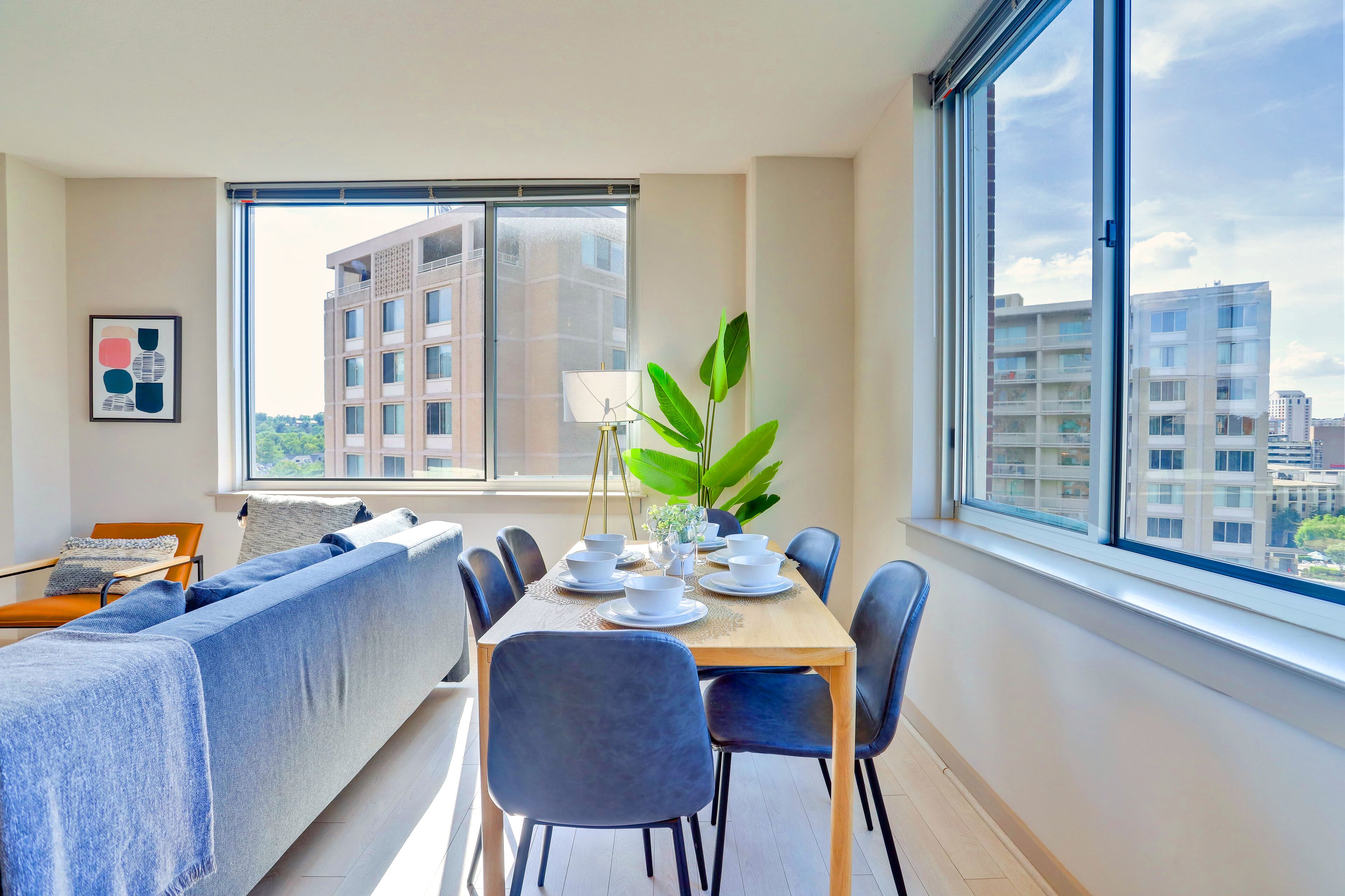 A modern living room with a sofa, dining table, chairs, and a large window offering a city view.