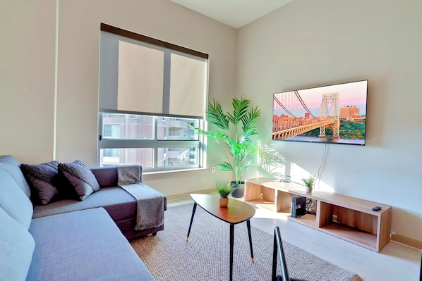 A modern living room with a grey sofa, a small coffee table, a TV mounted on the wall, and a large window with a cityscape view. - Crystal City Apartments near S Bell Street - We Host (Arlington)