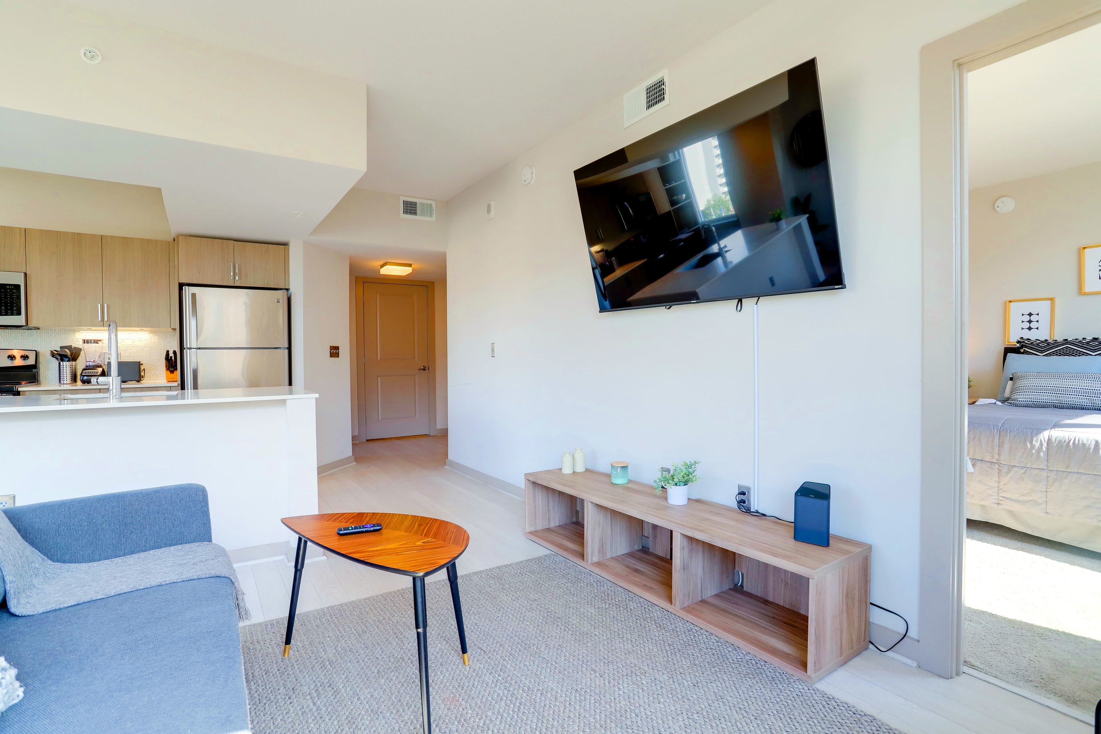 A modern living room with a sofa, a coffee table, a TV mounted on the wall, and a kitchen area in the background.