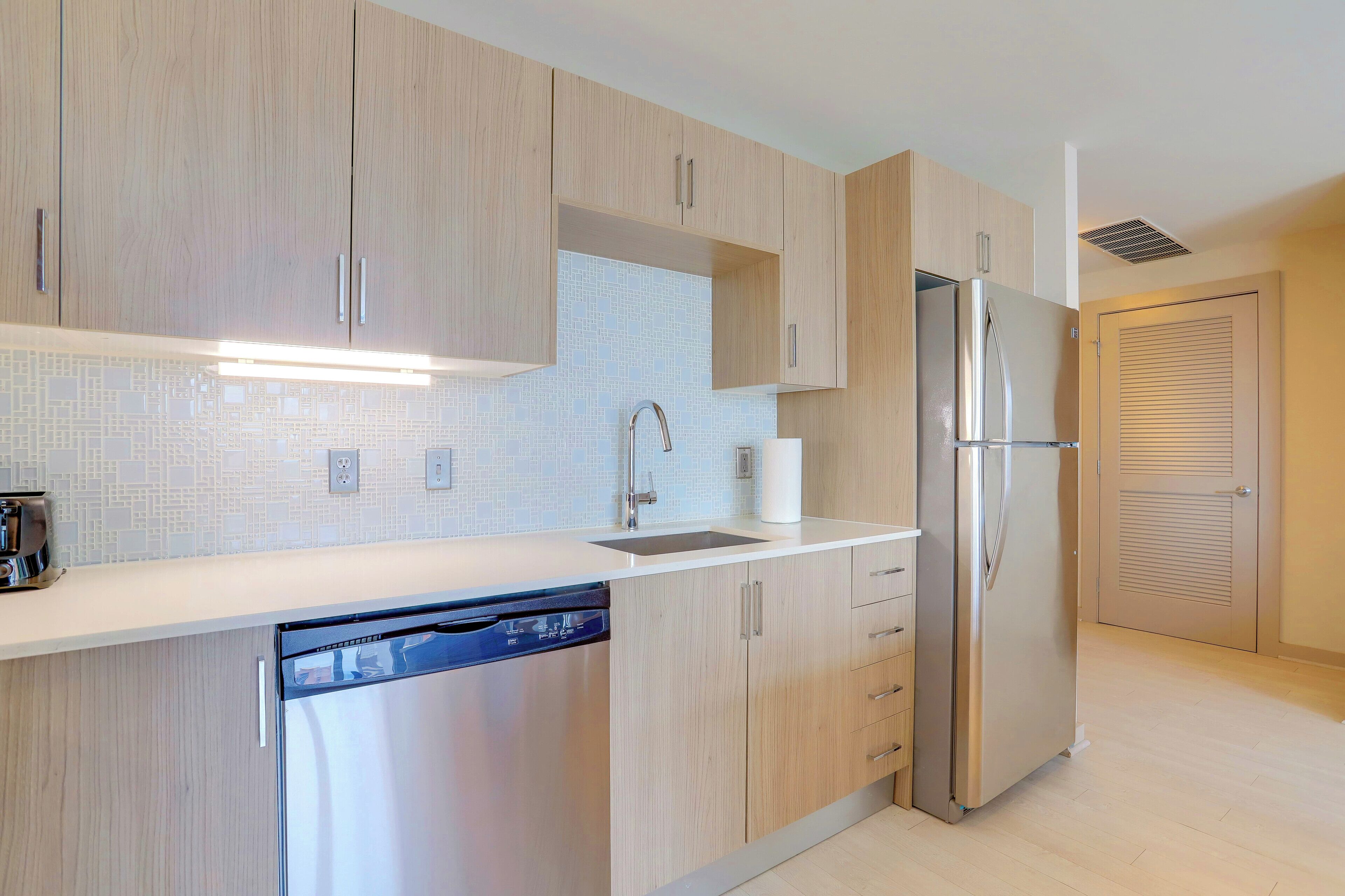 A modern kitchen with light wood cabinets, a stainless steel refrigerator, a dishwasher, and a tiled backsplash.