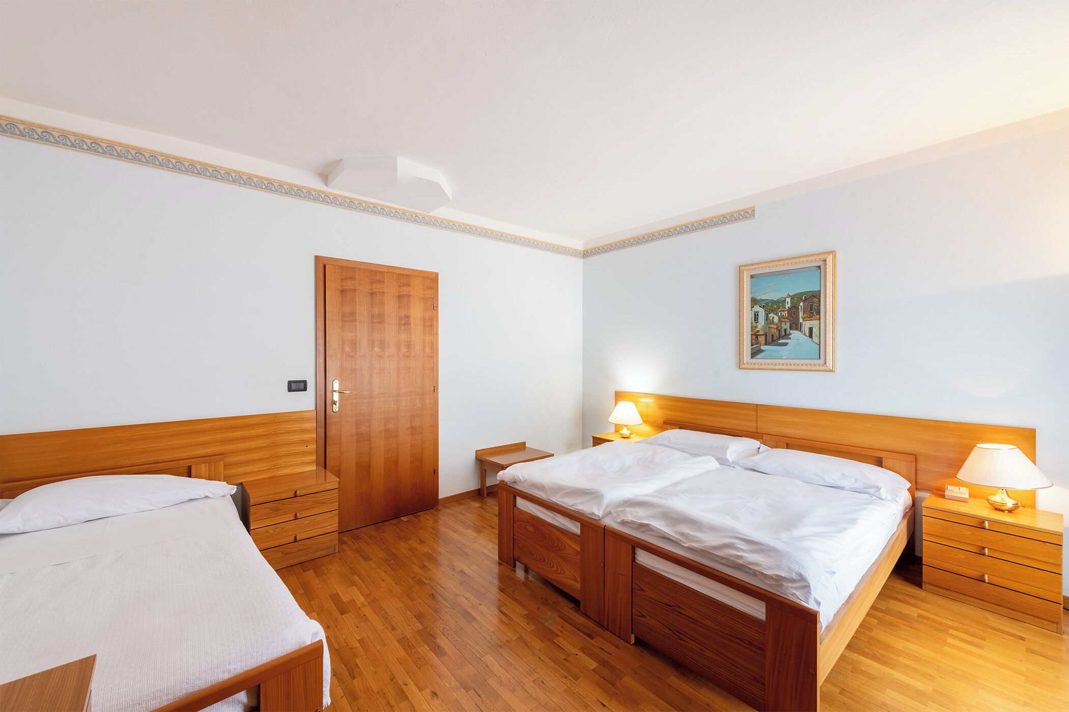 A hotel room with two wooden beds, a wooden wardrobe, a small bedside table, a nightstand with a lamp, and a framed picture on the wall.