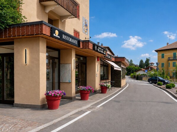 A street view with a building featuring a restaurant named 'Ristorante' and a bar named 'Albergo-Bar Villa Novina.' There are potted plants in front of the building. - VILLA NUOVA (Romeno)