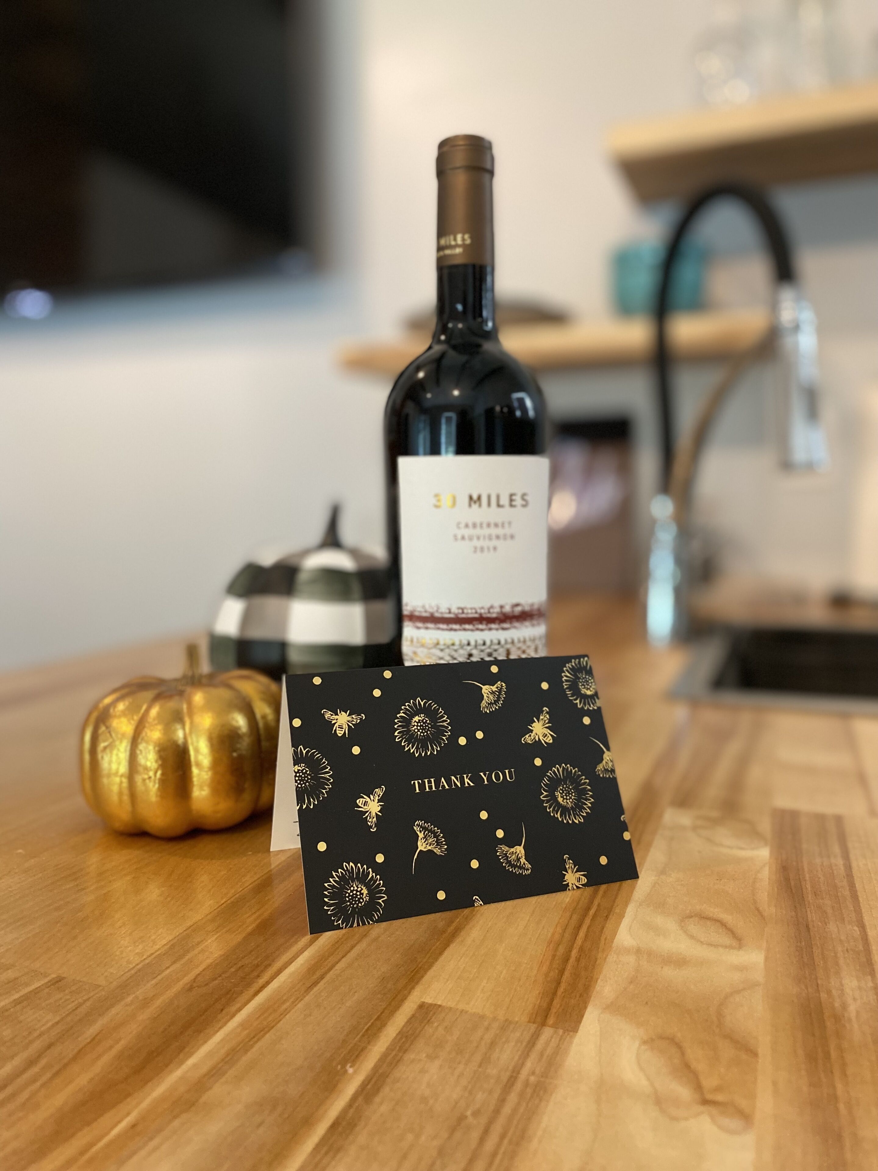 A bottle of wine labeled "30 Miles" on a wooden table with a black card featuring gold designs and the text "THANK YOU".