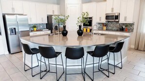 A modern kitchen with a central island, white cabinetry, stainless steel appliances, and black bar stools. - Gorgeous 13BR Encore Resort Next to Disney - 348 (Kissimmee)