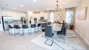 A modern kitchen with white cabinets, a central island with bar stools, and a dining area with a marble rug. - Gorgeous 13BR Encore Resort Next to Disney - 348 (Kissimmee)