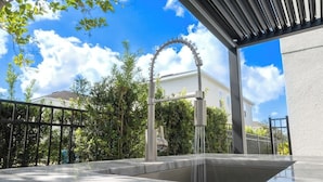 Una cocina exterior moderna con pileta y grifo de acero inoxidable, rodeada de follaje y un cielo azul despejado.