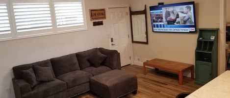 Ein Wohnzimmer mit Ecksofa, einer Holzbank, einem an der Wand befestigten Fernseher und einer Tür mit einem Schild darüber.