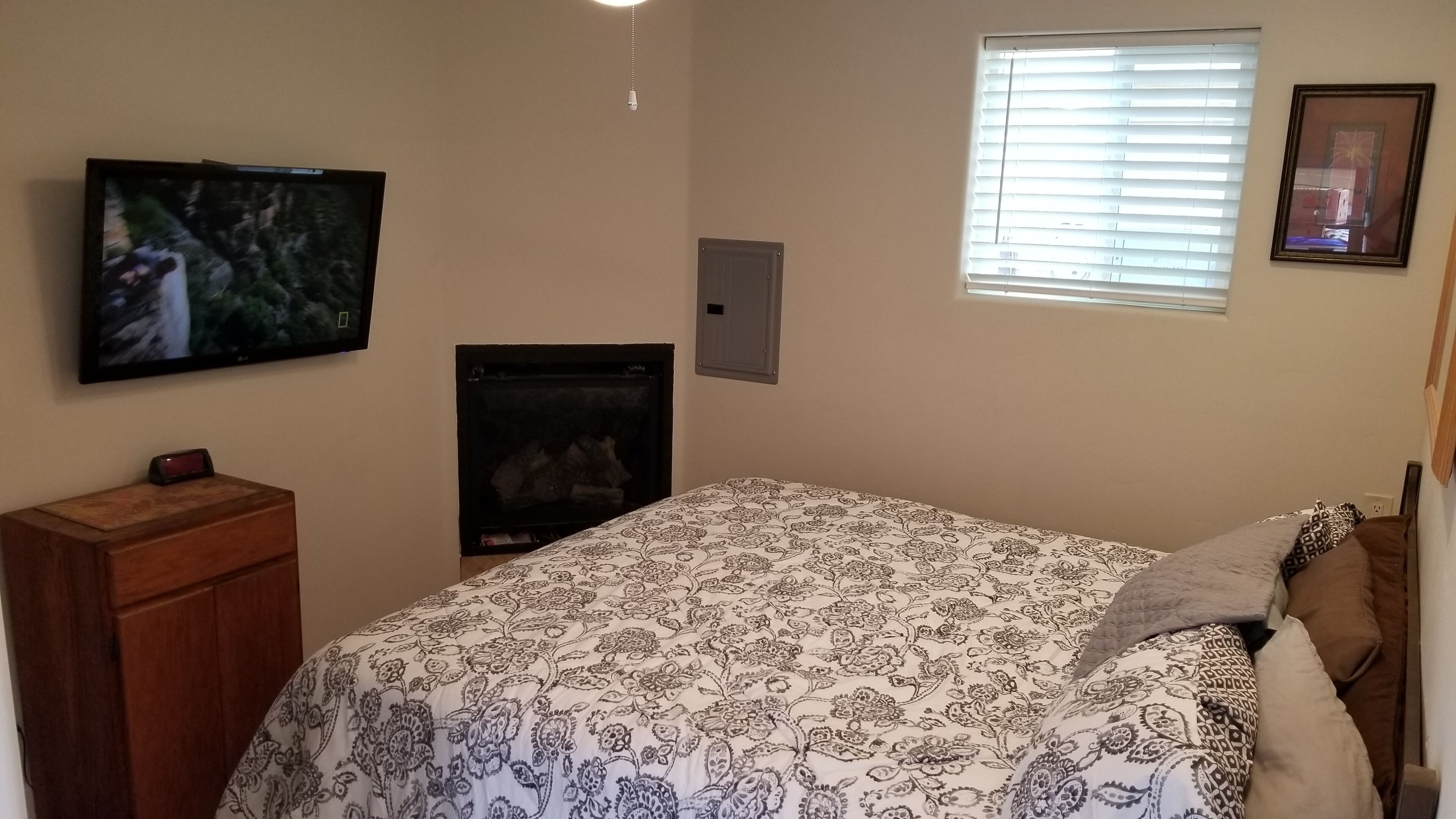 A bedroom with a bed, a TV mounted on the wall, a fireplace, and a window with blinds.