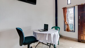 Une petite pièce avec une table, deux chaises, un ordinateur portable, un téléviseur fixé au mur, une fenêtre avec des rideaux et une porte.