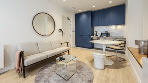 Una sala moderna con un sofá, una mesa de centro y un espejo redondo. Se observa una zona de cocina con armarios azules y una mesa de comedor con sillas.