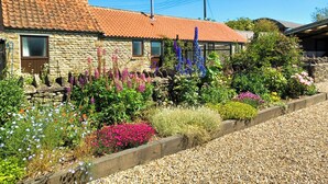 A stone house with a tiled roof, surrounded by a gravel driveway and a variety of flowering plants. - 2 bedroom accommodation in Levisham, near Pickering (Levisham, near Pickering)