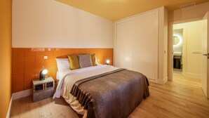 Une chambre à coucher comprenant un lit, des tables de chevet, une lampe fixée au mur et une porte donnant accès à une salle de bain.