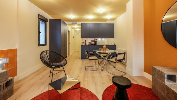 Une cuisine moderne avec des meubles de couleur sombre, un coin repas avec une table en verre et des chaises en métal, et un tapis rouge sur le sol.
