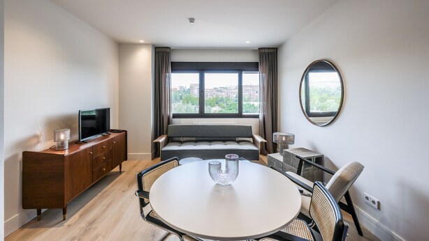 Una habitación de hotel moderna con un ventanal grande, un sofá, una mesa de comedor y un televisor sobre un mueble de madera.