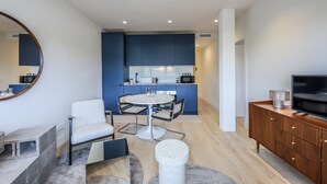 Una cocina moderna con armarios azules, una zona de comedor con una mesa redonda y sillas, un televisor sobre un mueble de madera y un espejo grande en la pared.