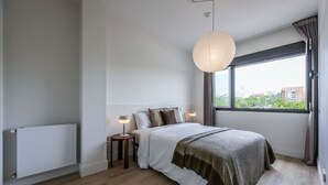 Un dormitorio con una cama grande, un radiador, una ventana con vistas a edificios y una lámpara colgante.