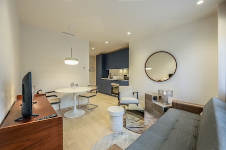 A modern living room with a grey sofa, a round mirror, a dining table with chairs, and a TV on a wooden desk.